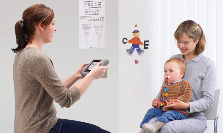 A woman is holding a baby in front of a white wall, seeking the best vision test near me.