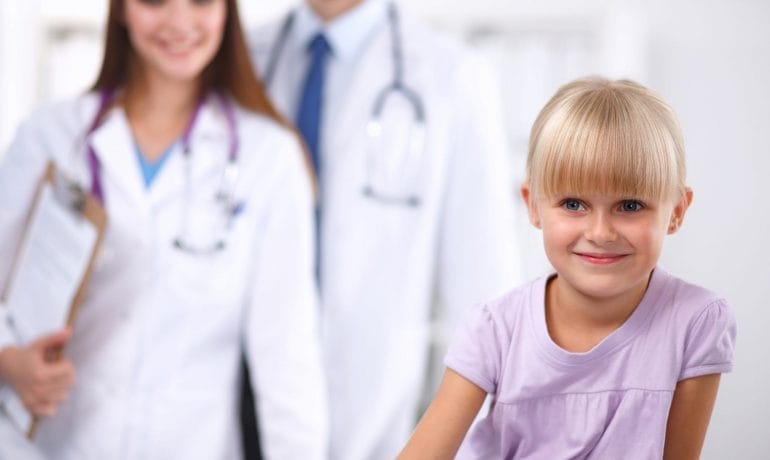 A young girl smiling in the foreground with two healthcare professionals standing in the background.
