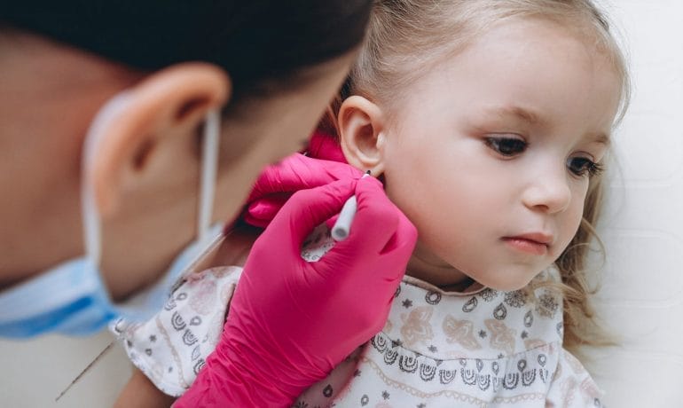 A young child is getting their ear pierced by a professional wearing pink gloves and a face mask at an asthma care center in Sugar Land. The child looks calm and is wearing a printed shirt.