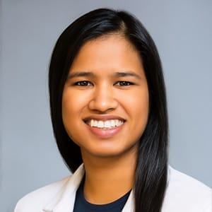 Doctor Pillai A Pediatrician Sugar Land with straight black hair, wearing a white coat and dark shirt, smiles warmly in front of a plain gray background.